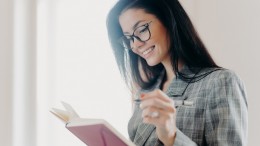 Sideways shot of pleasant looking brunette woman has happy expression makes notes in planner, does remote job, creats planning, wears optical eyeglasses and formal clothes, poses near window